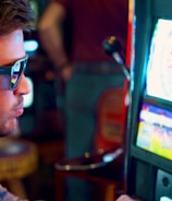 A man wearing sunglasses is intently focused on a slot machine, with colorful lights and graphics illuminating the screen. The scene is set indoors, likely in a casino or gaming area. His attire includes a patterned shirt and a blazer.