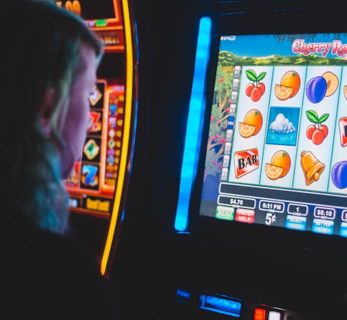 A person is closely observing a digital slot machine with colorful fruit symbols, bars, and a bell. The slot machine screen displays symbols like oranges, plums, cherries, and clouds. The background is dimly lit, creating a focused and somewhat intense atmosphere.