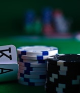 A casino scene featuring two playing cards, the ace and king of clubs, partially concealed by a player's hand. Nearby, there are stacks of multicolored poker chips on a green felt table, with blurred colorful background elements suggesting other chip stacks or cards.
