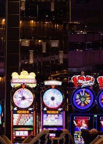 A lively casino scene featuring a row of bright, colorful slot machines with themes like Monopoly, Michael Jackson, and Lord of the Rings. The gaming machines display a variety of flashing lights and digital screens, creating an energetic and exciting atmosphere. Reflections of the lights are visible in glossy surfaces above the machines.
