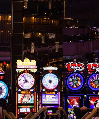 A lively casino scene featuring a row of bright, colorful slot machines with themes like Monopoly, Michael Jackson, and Lord of the Rings. The gaming machines display a variety of flashing lights and digital screens, creating an energetic and exciting atmosphere. Reflections of the lights are visible in glossy surfaces above the machines.
