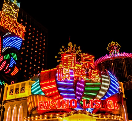 Bright neon lights and colorful signage adorn the exterior of Casino Lisboa, creating a vibrant and lively atmosphere. The façade features bold, multicolored designs with a mix of English and Chinese characters prominently displayed. The lights form intricate patterns and shapes, contributing to the energetic ambiance of the scene.